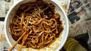 A bowl of jajangmyeon recipe noodles with thick black bean sauce, partially eaten, sits on a table with wooden chopsticks and Korean newspapers and pickled radish slices in the background.